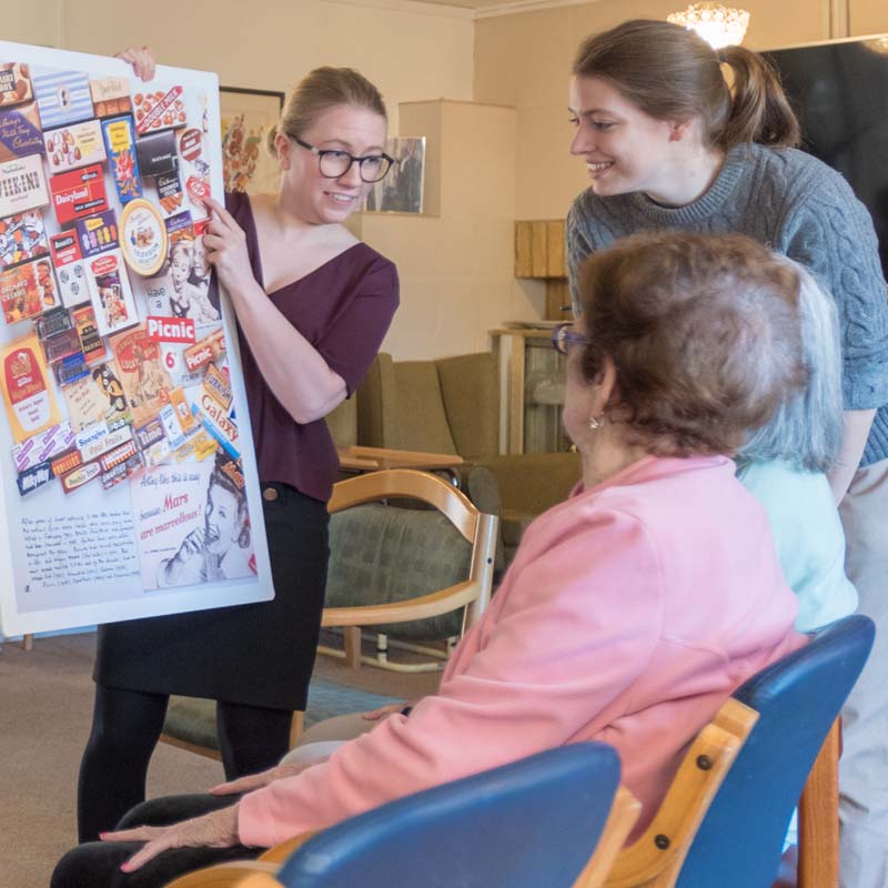 Four people inside a care home. One is standing up and holds a big collage on paper of famous chocolate brands. They are young and wearing a red top and black trousers. The person next to them is young and smiling while looking at the brands. There are two older ladies sitting on the chairs in between them facing the collage of chocolate brands.