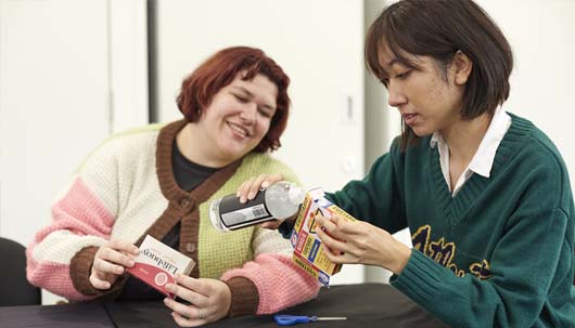Two women sat next to each other on a table. One has red hair and a striped cardigan and is holding a red soap packet from the brand lifebuoy. The other has black hair, a green jumper and is holding a red, yellow and blue striped custard packet from the brand birds eye.