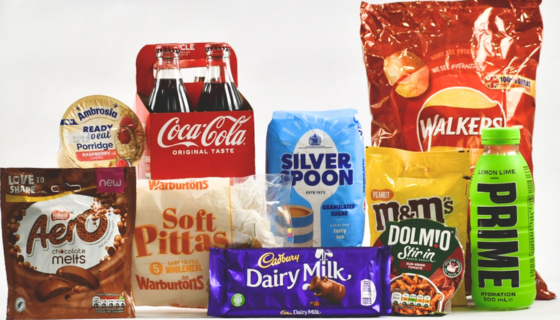 A selection of popular grocery brands on a white background, including a purple Cadbury Dairy Milk bar, a green bottle Prime energy drink, and a large red packet of Walkers crisps
