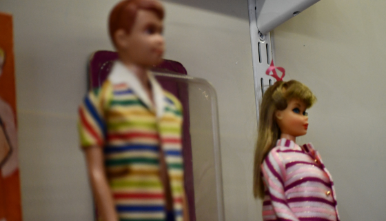 Two dolls on a shelf: the left has short red hair and a multicolored striped shirt; the right has long blonde hair and a pink and white striped jacket.