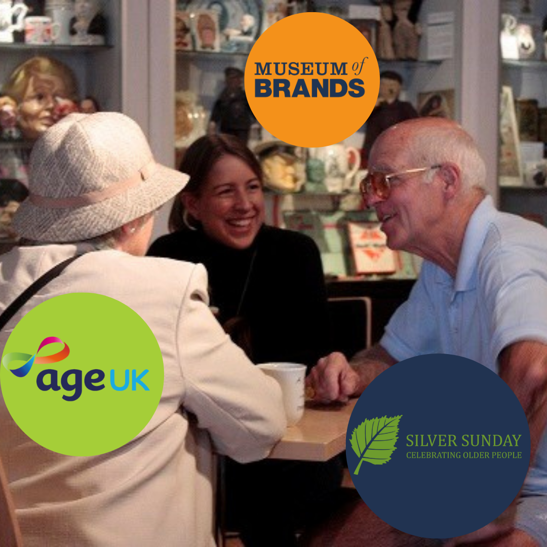 Three people sat around a museum cafe table. There is a display of bottles in the background. In the foreground are two older people - a woman in a white hat and jacket and a man in a light blue shirt with glasses. There is a younger woman with brown hair and a black jacket sitting on the other side of the table and laughing with them.